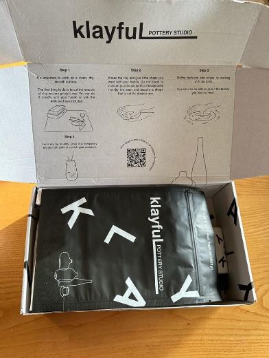 Offene Verpackung des Klayful Pottery Studios mit Anleitung im Deckel und einem Blick auf die verpackten Inhalte, einschließlich eines schwarzen Aprons mit dem Klayful Logo