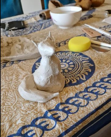 Unbemalte, lufttrocknende Tonfigur eines Fuchs auf einem gemusterten Tischtuch, neben einer Schale und einem gelben Schwamm