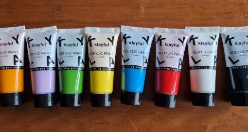 Verschiedene Farbtuben des Klayful Acrylfarbensets auf einem Holztisch, sortiert nach Farbe – Orange, Weiß, Violett, Grün, Gelb, Blau, Rot und Schwarz