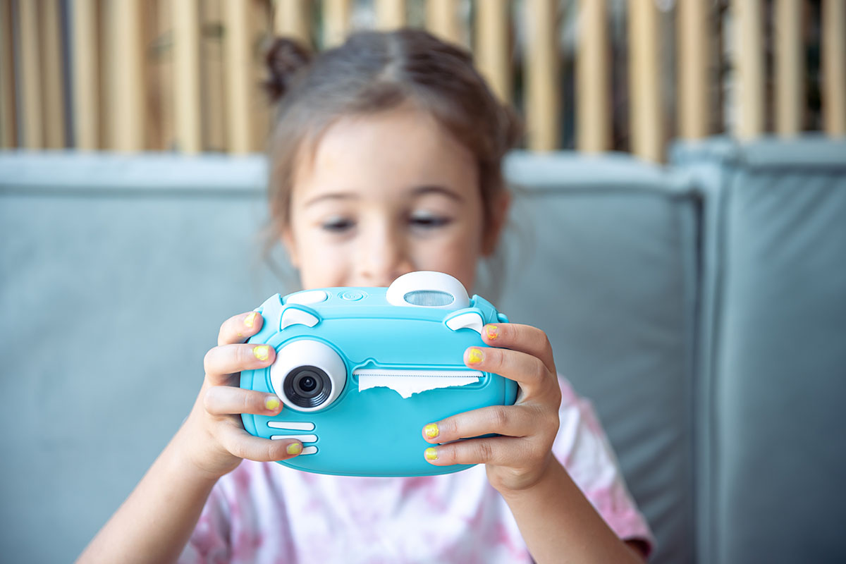 Ein kleines Mädchen hält eine blaue digitale Spielzeugkamera für Kinder für den Sofortdruck von Fotos in ihren Händen.